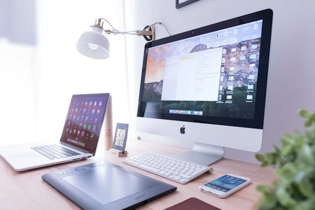 Trends in Responsive Web Design Apple Monitor Screen Show Responsive Web Design stand on Wooden Table with White color keyboard and Laptop, iPhone, Black color Touch pad, White color Table Lamp at corner green plant.
